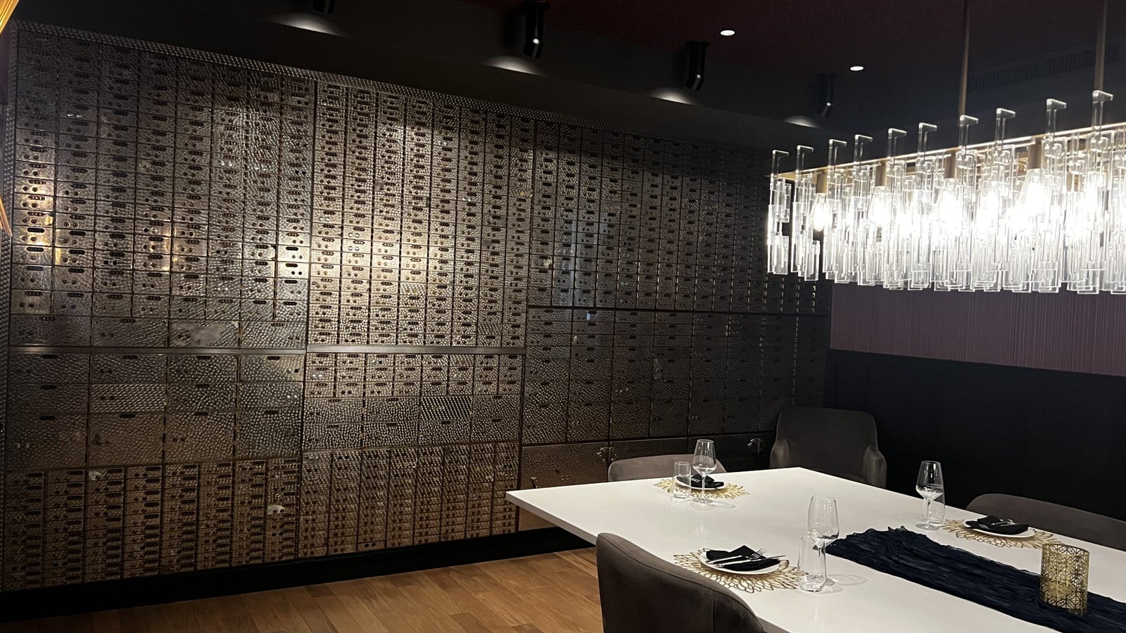 Interior dining room with a wall of vintage bank safe deposit boxes and a modern chandelier above a set dinner table.
