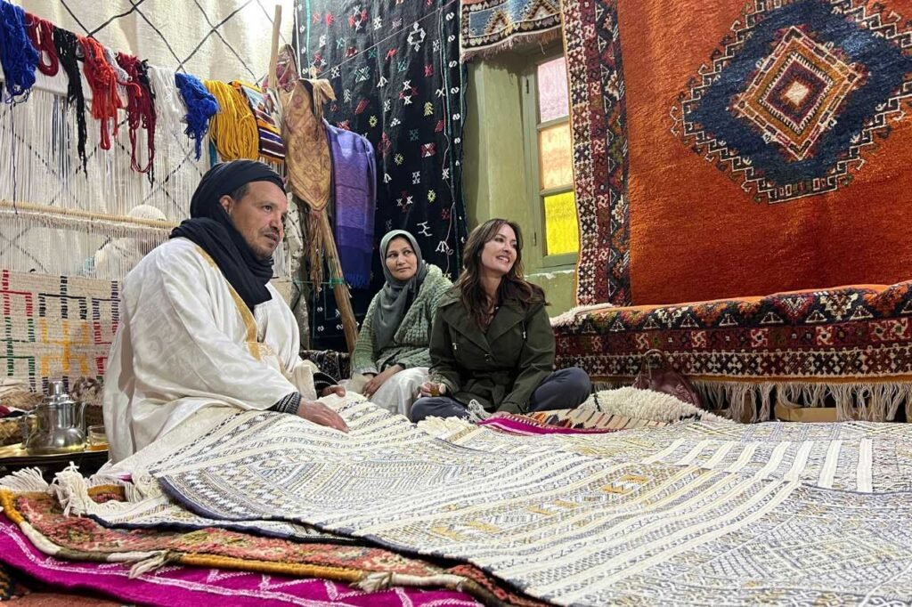 A Berber carpet maker and his wife showcase traditional handwoven rugs inside their workshop in Morocco, sharing the craft with a visitor seated among colorful textiles.