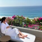 Guest relaxing on a balcony at Grand Velas Boutique Los Cabos, sipping coffee in a white robe with vibrant pink bougainvillea and ocean views in the background.