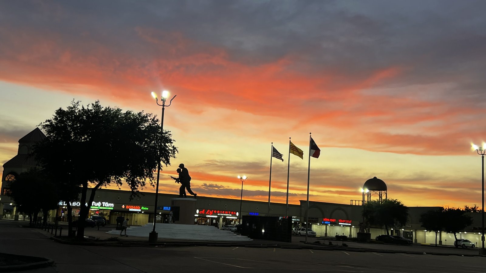 Sunset over Houston Asiatown on Bellaire Boulevard, with vibrant orange and pink skies framing the silhouette of the Vietnam War Memorial statue, multiple national flags, and local shops, capturing the blend of crawfish culture and Little Saigon’s vibrant community.