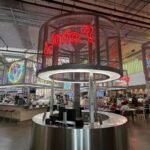 Interior of Post Houston food hall featuring neon signs, modern industrial design, and a central circular info desk surrounded by eateries and seating areas.