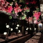 Elegant restaurant interior with white tablecloth dining tables, globe pendant lamps, and a dramatic ceiling installation of lush greenery and cascading pink, red, and purple flowers - experiencing culinary culture in Bayou City.
