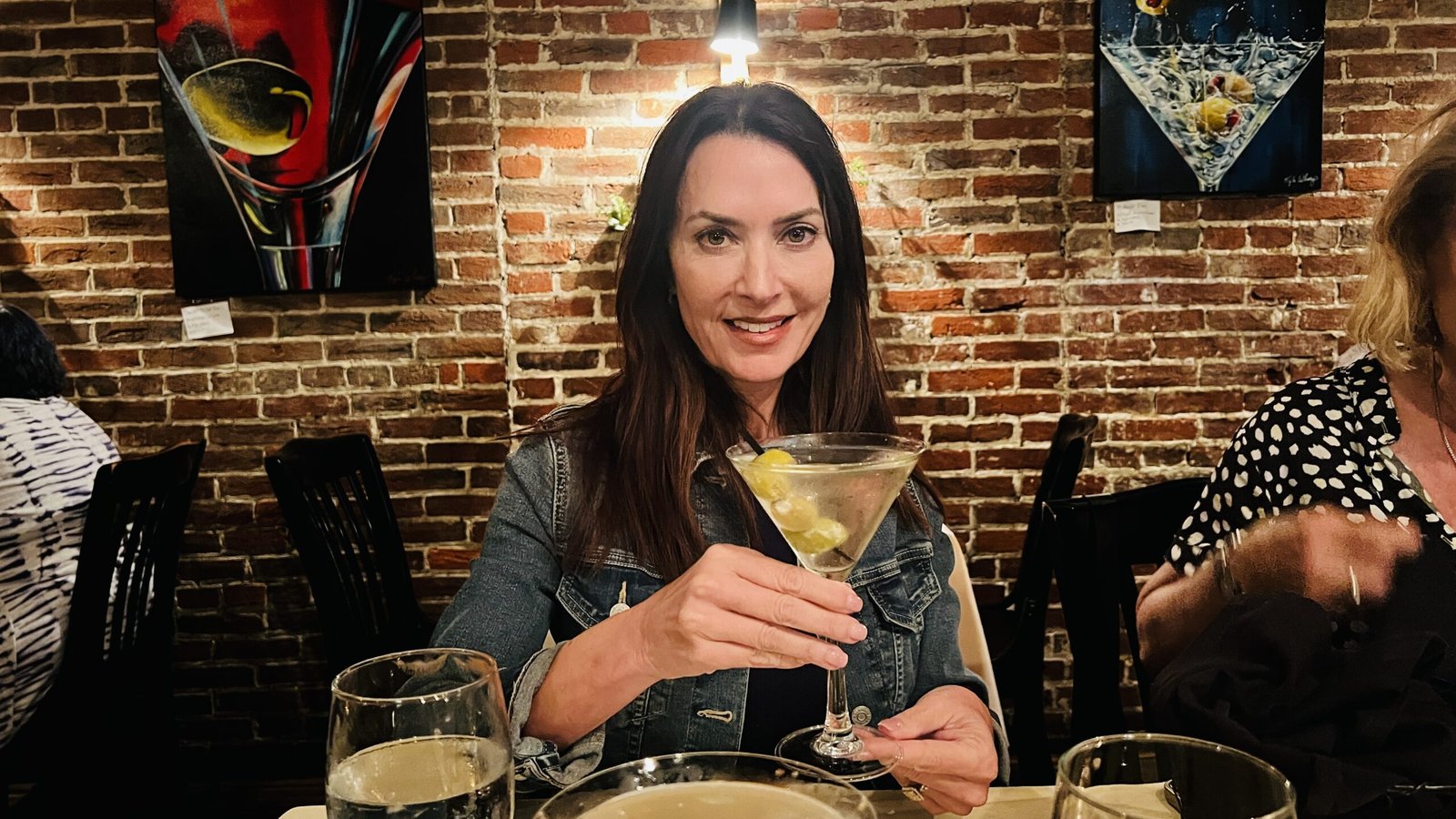 Woman smiling while holding a martini glass inside a cozy brick-walled restaurant.
