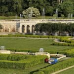 A beautifully landscaped garden with neatly trimmed hedges, fountains, and a wide green lawn in front of an ornate stone terrace at Longwood Gardens, Pennsylvania. A couple of chairs and a picnic blanket are set up on the grass, adding a touch of leisure to the grand setting.
