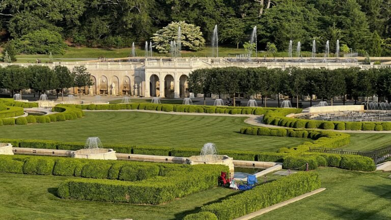 A beautifully landscaped garden with neatly trimmed hedges, fountains, and a wide green lawn in front of an ornate stone terrace at Longwood Gardens, Pennsylvania. A couple of chairs and a picnic blanket are set up on the grass, adding a touch of leisure to the grand setting.