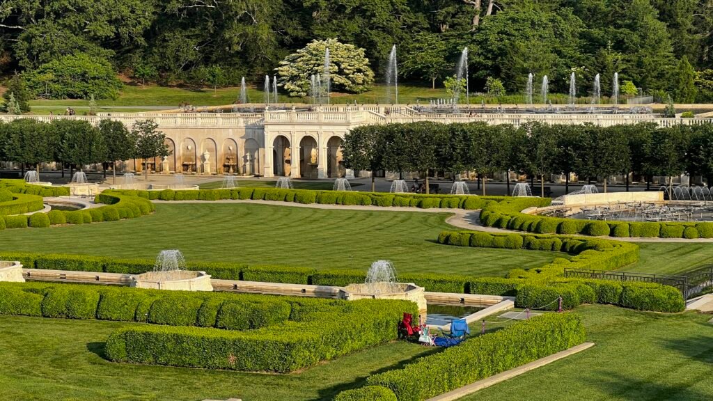 A beautifully landscaped garden with neatly trimmed hedges, fountains, and a wide green lawn in front of an ornate stone terrace at Longwood Gardens, Pennsylvania. A couple of chairs and a picnic blanket are set up on the grass, adding a touch of leisure to the grand setting.