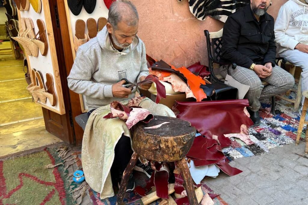 A Moroccan leather artisan cutting dyed leather pieces by hand at a souk workshop, surrounded by vibrant scraps and handmade slippers.