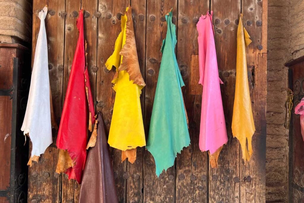 Brightly dyed leather hides in red, yellow, turquoise, pink, white, and brown hanging to dry on a rustic wooden wall in Marrakech’s tanneries.