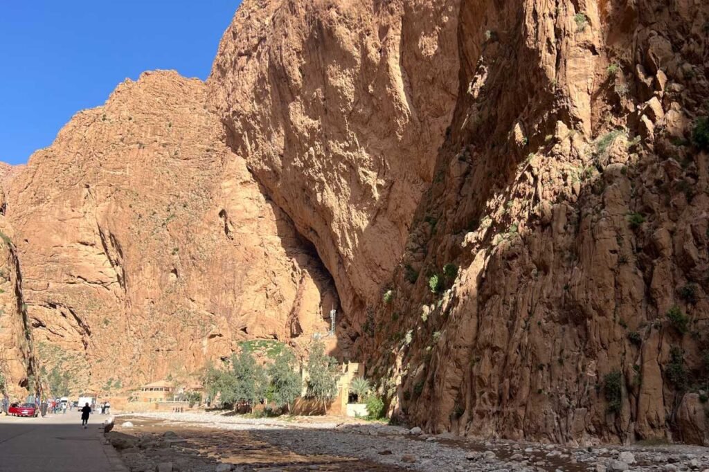 Towering red limestone cliffs of Todra Gorge in Morocco with a narrow passage and scattered greenery at the base under a clear blue sky.