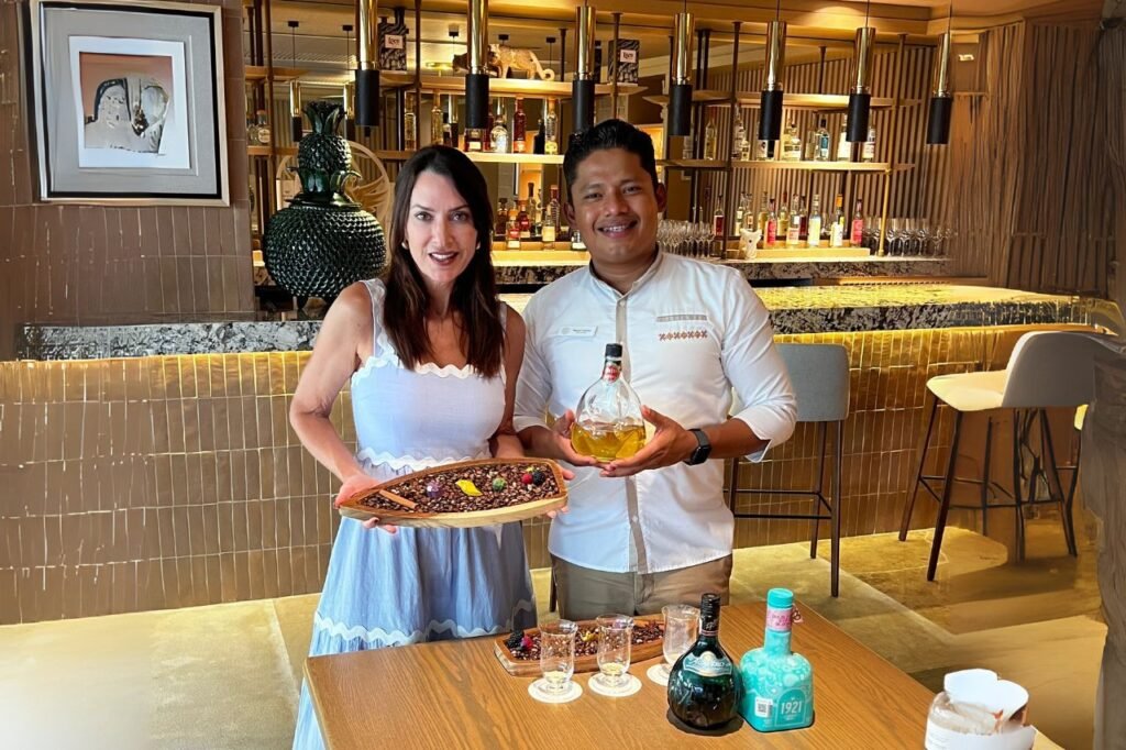 Two people standing in a stylish bar holding a tray of cacao beans and a bottle of liquor, with tasting glasses and bottles on the table in front of them.