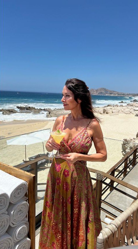 Woman in a patterned sundress holding a cocktail, standing on a beachside deck with ocean waves and sandy shore in the background.
