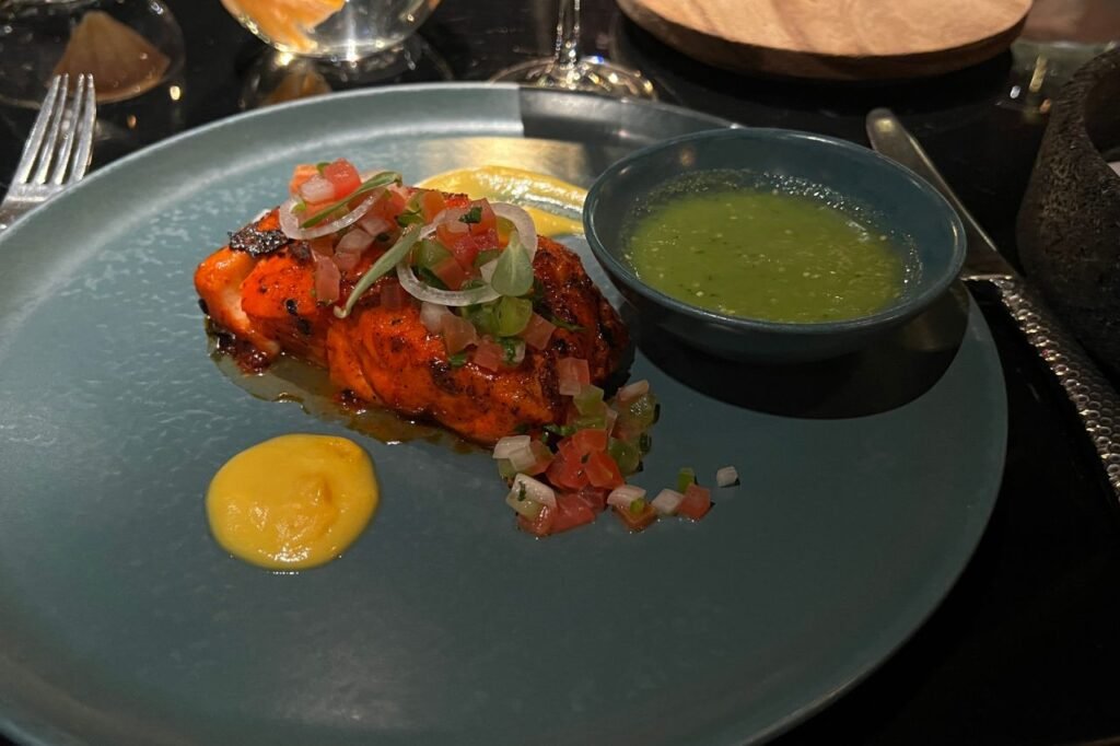 Grilled salmon topped with fresh pico de gallo and onions, served with yellow sauce and a side of green salsa on a dark ceramic plate.