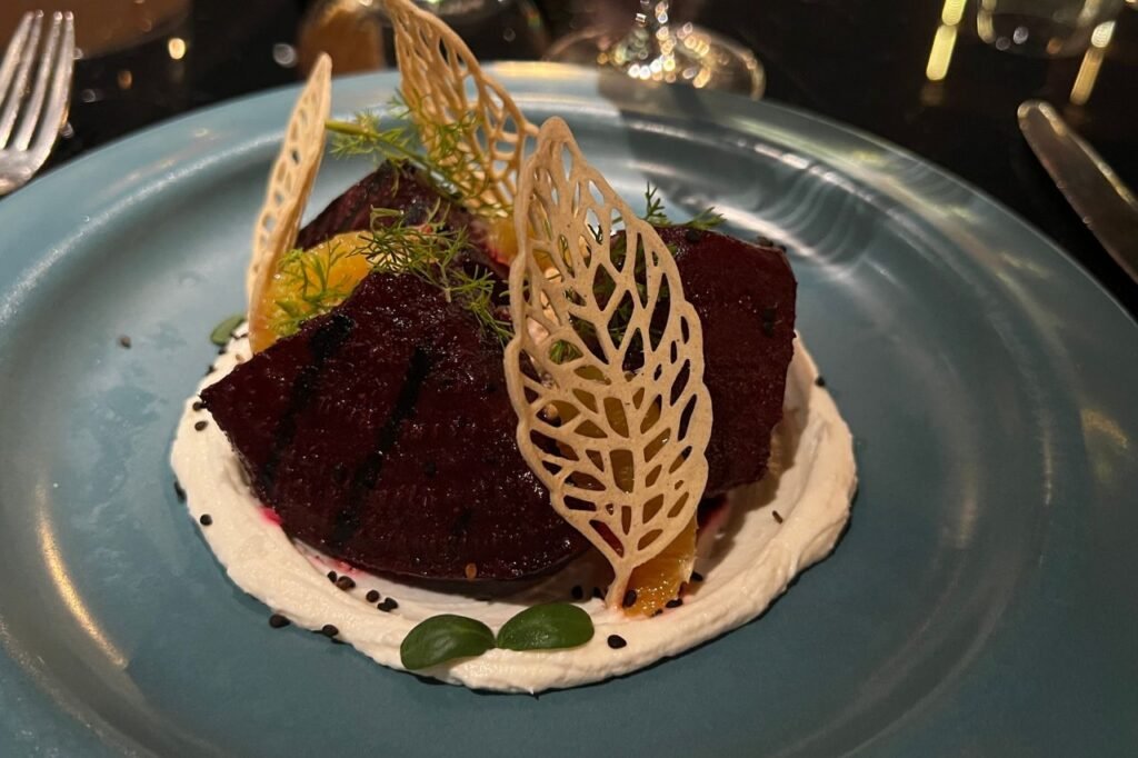 Plated gourmet dish with grilled beets on a bed of creamy sauce, garnished with orange slices, herbs, and delicate crispy leaf-shaped toppings.