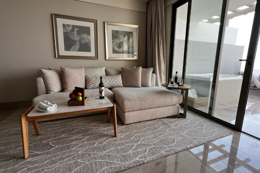 Elegant suite interior at Grand Velas Boutique Los Cabos featuring a cozy beige sectional sofa with plush pillows, a coffee table with wine and fresh fruit, and a private terrace with a jacuzzi visible through sliding glass doors.