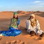 A woman in a vibrant blue and green dress and a Berber guide in traditional white robes sit on the golden sand dunes of the Sahara Desert during sunset in Morocco