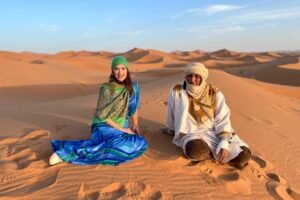 A woman in a vibrant blue and green dress and a Berber guide in traditional white robes sit on the golden sand dunes of the Sahara Desert during sunset in Morocco