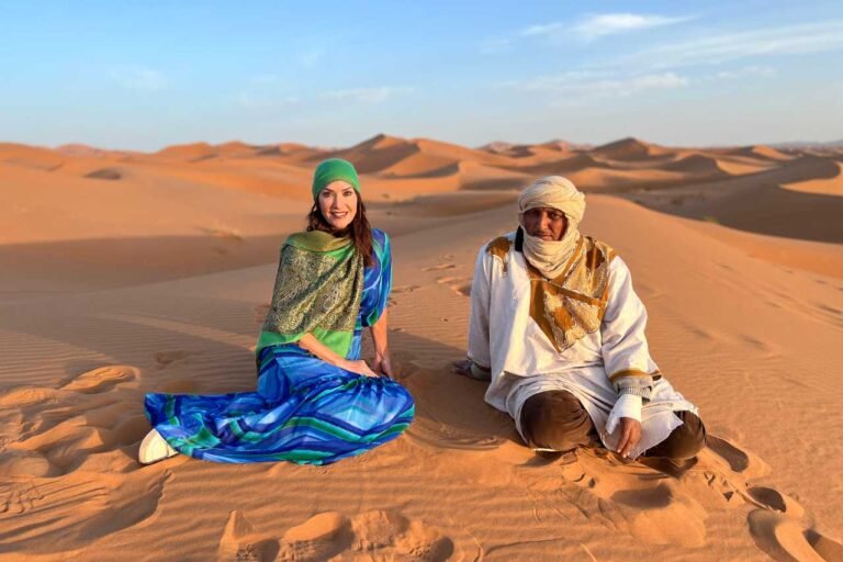 A woman in a vibrant blue and green dress and a Berber guide in traditional white robes sit on the golden sand dunes of the Sahara Desert during sunset in Morocco