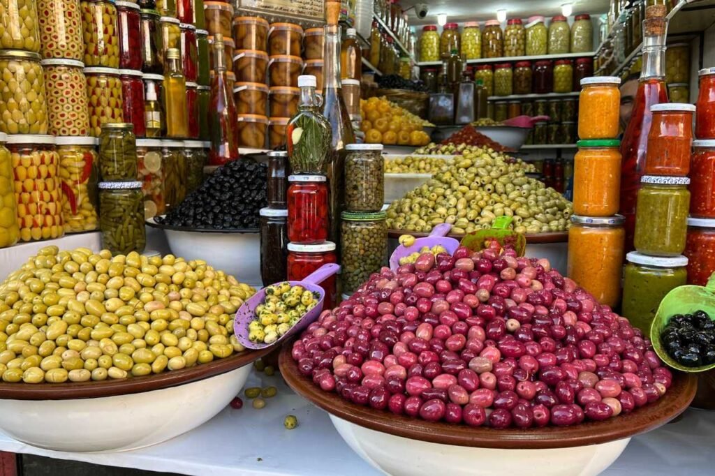 Colorful display of Moroccan olives and pickled vegetables in a Marrakech market, with jars of preserved lemons, peppers, and spices stacked behind vibrant bowls of green, black, and red olives.
