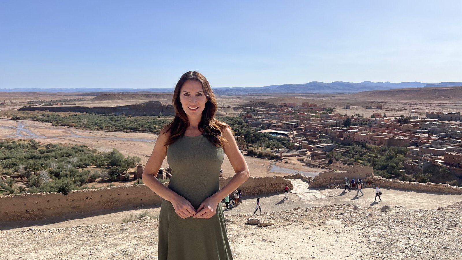 Woman in green dress standing on a hilltop overlooking Aït Benhaddou and the surrounding desert valley.
