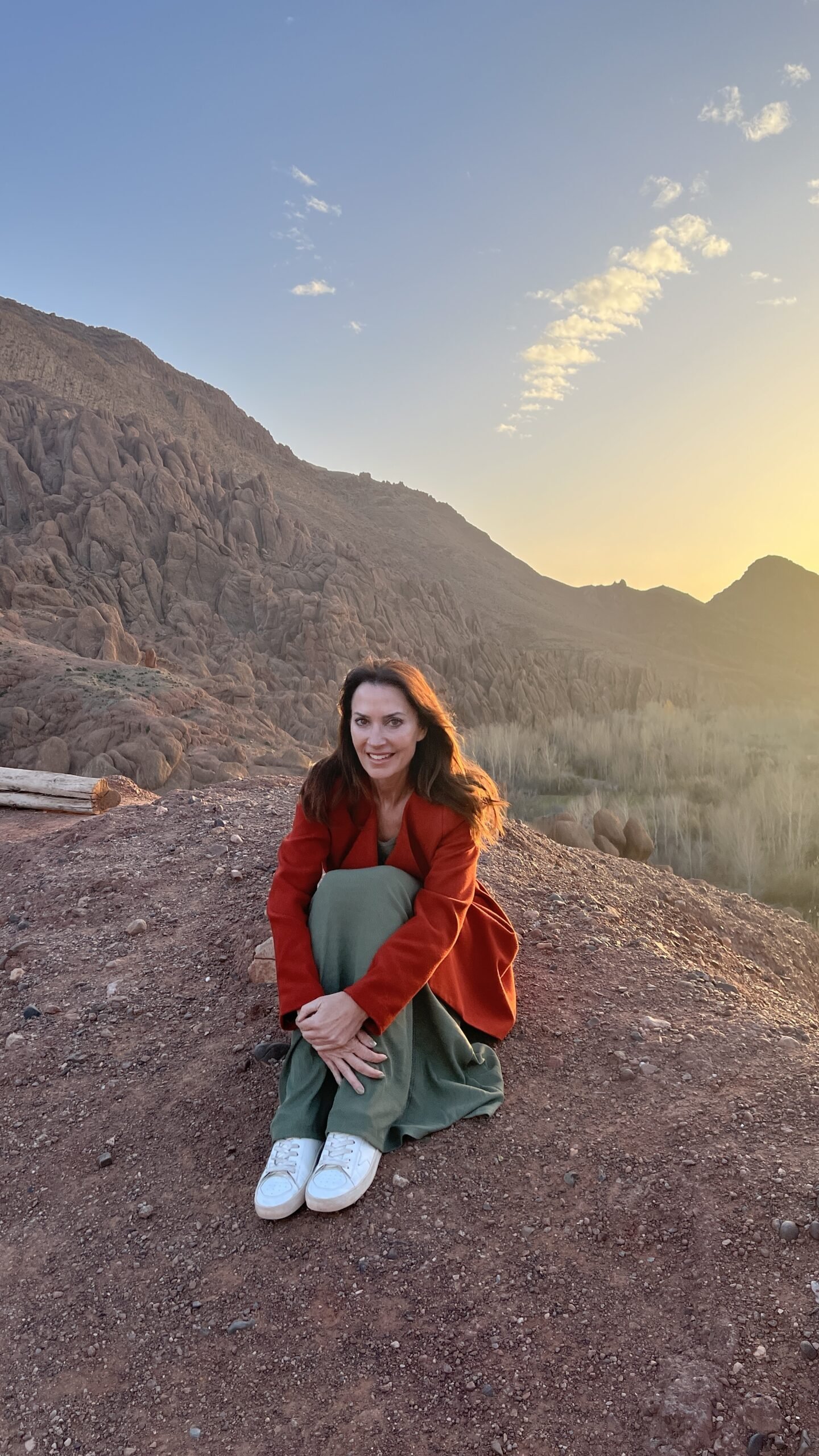 Woman sitting on rocky ground at sunset with the High Atlas Mountains rising behind her.