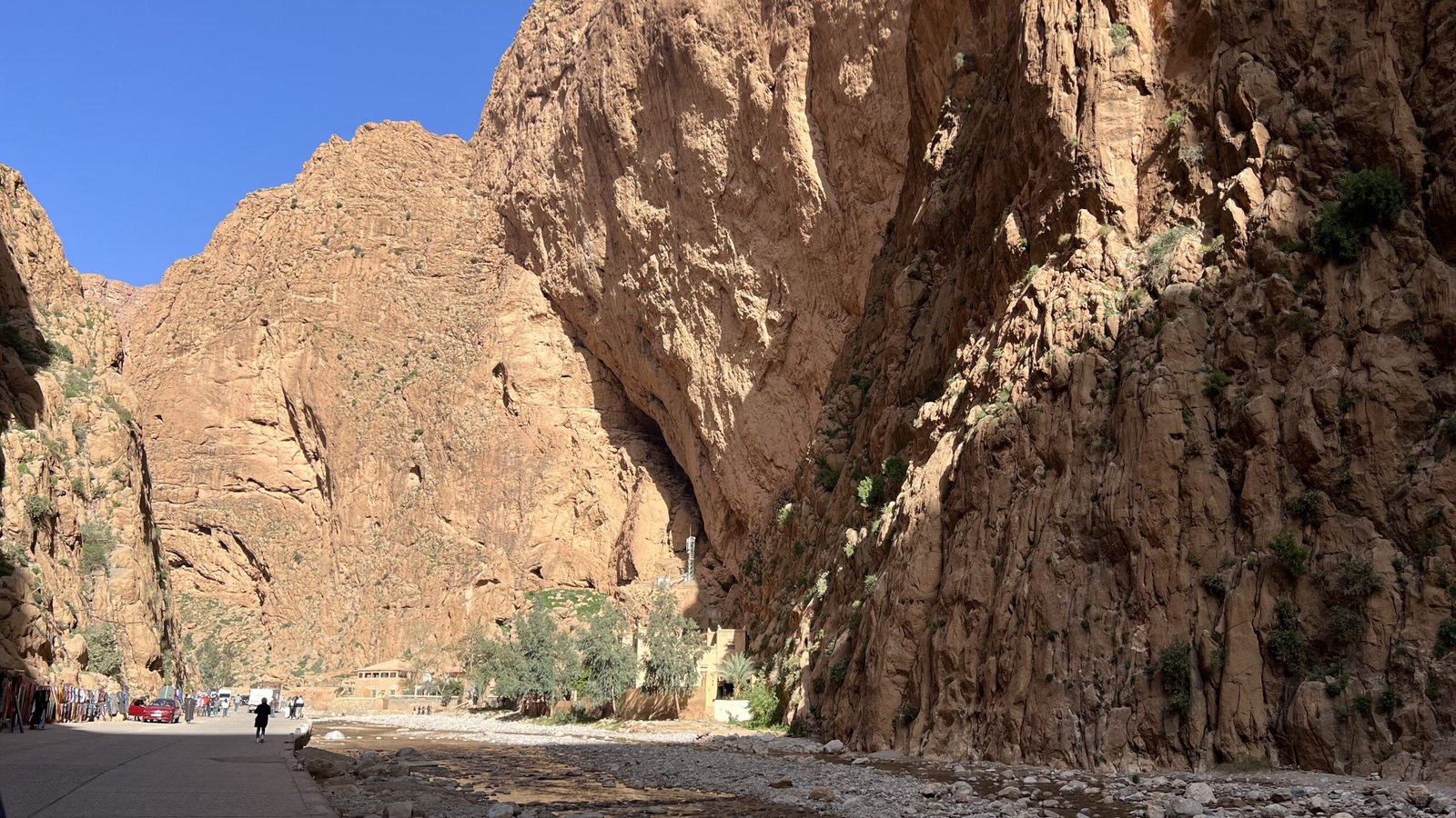 Road running through towering red cliffs of Dades Gorge in Morocco with cars and trees below.