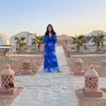 Woman in a blue patterned dress standing on a walkway lined with lanterns at a Sahara desert camp surrounded by white tents and palm trees.