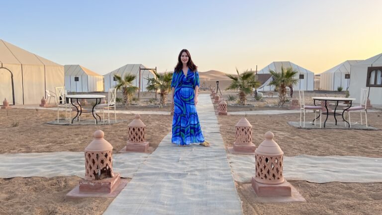 Woman in a blue patterned dress standing on a walkway lined with lanterns at a Sahara desert camp surrounded by white tents and palm trees.