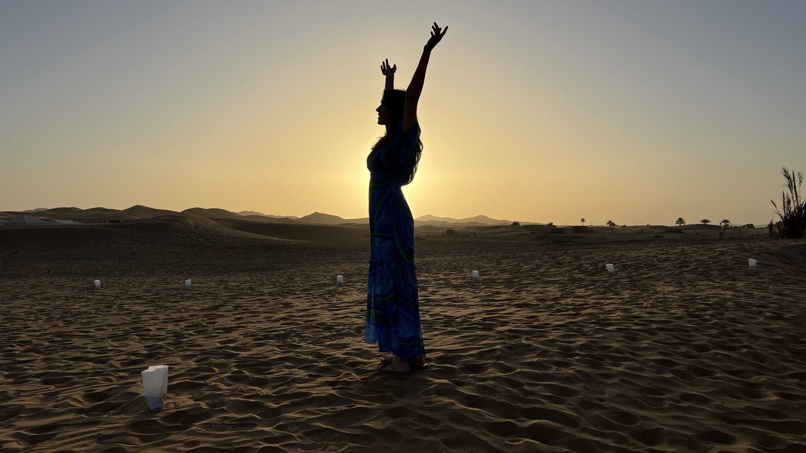 Woman in a blue dress raising her arms at sunrise on the dunes of Erg Chebbi in the Sahara Desert.