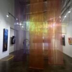 Colorful sheer fabric panels hang in the center of a contemporary art gallery at Untitled Art Houston, with abstract paintings displayed on white walls and spotlights illuminating the space.