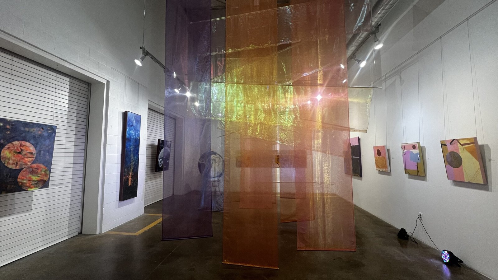 Colorful sheer fabric panels hang in the center of a contemporary art gallery at Untitled Art Houston, with abstract paintings displayed on white walls and spotlights illuminating the space.