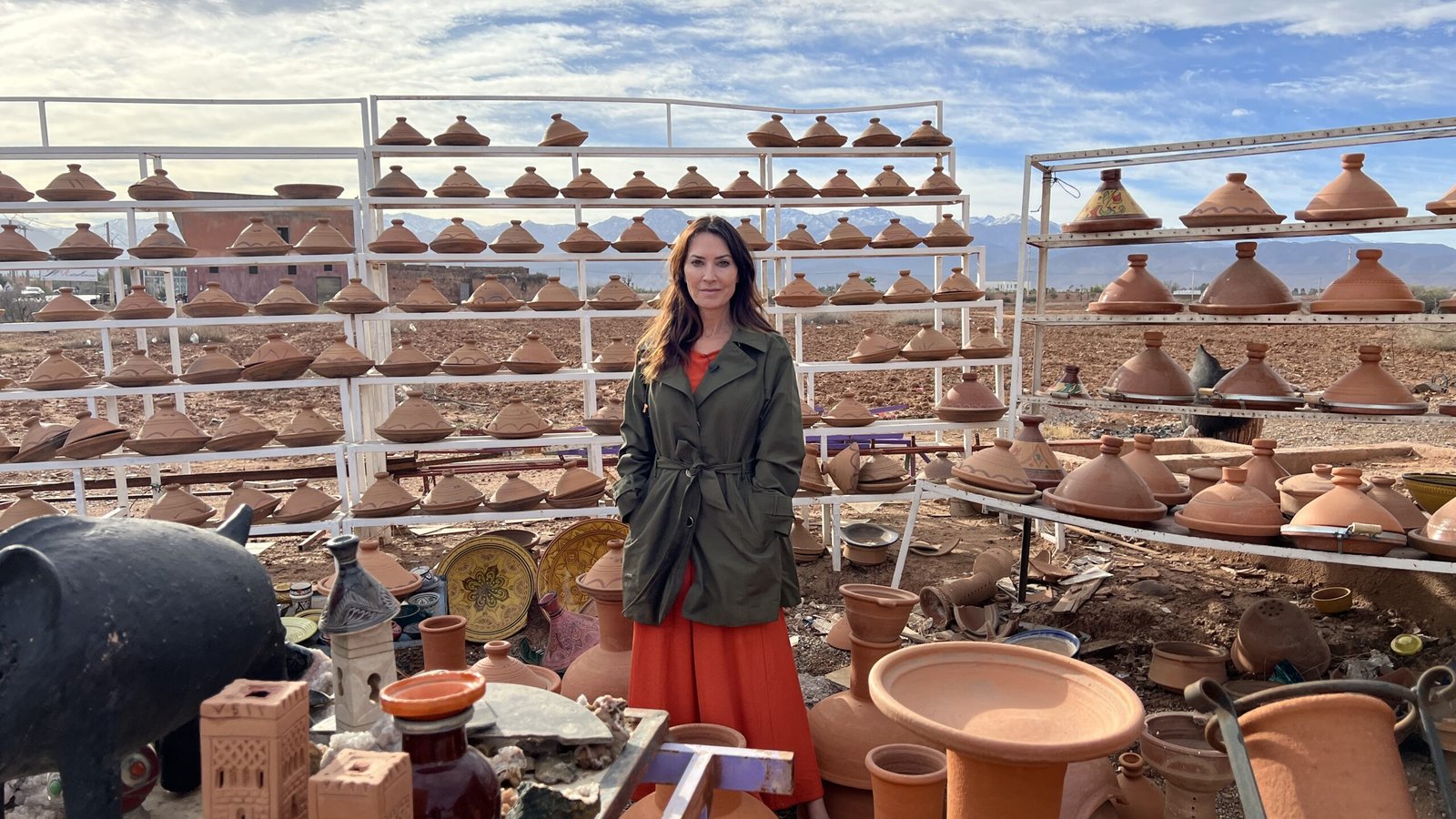 Woman in a red dress and green jacket standing among rows of clay tagines and pottery in an outdoor Moroccan market.