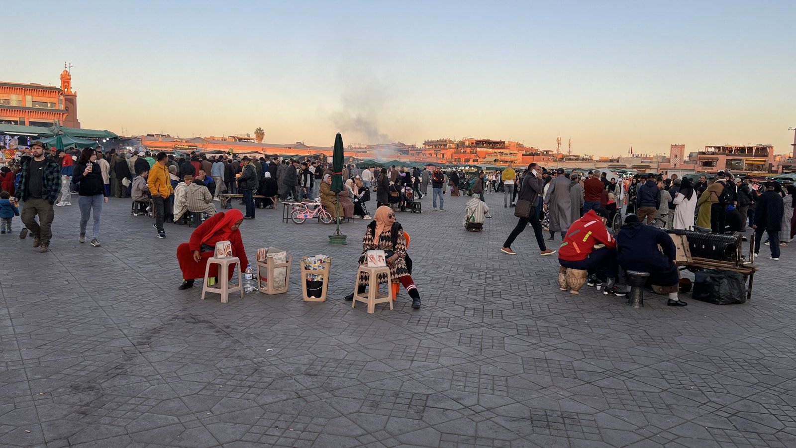 rowd gathered in Jemaa el-Fnaa square at sunset with vendors, smoke, and city backdrop.