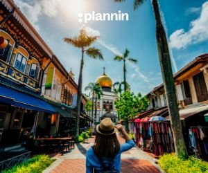 Traveler walking through a vibrant street lined with shops and palm trees, facing a golden-domed mosque under a sunny blue sky with the text “plannin” above.