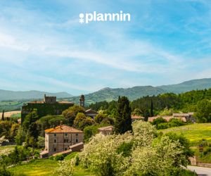 Scenic countryside village surrounded by green hills, trees, and historic stone buildings under a bright blue sky with the text “plannin” above.