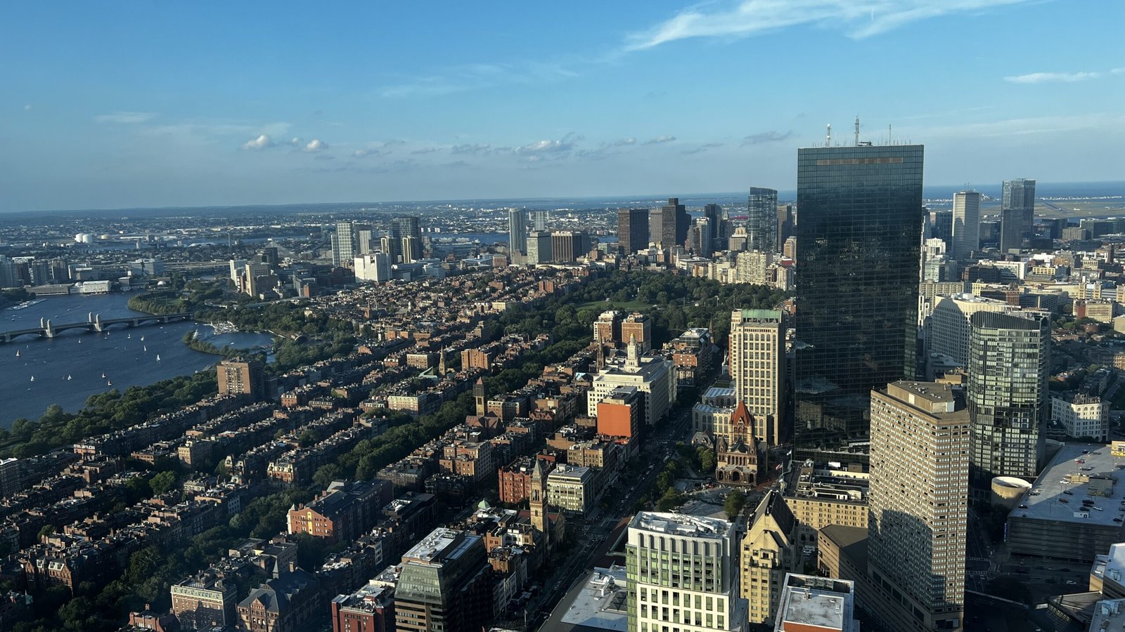 Aerial view of Boston cityscape from the Prudential Tower observation deck, showing downtown buildings and the Charles River under a blue sky.