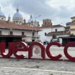 A large red “Cuenca” sign in an open plaza with a woman standing by the last letter. In the background are colonial-style buildings with terracotta roofs and the iconic blue-and-white domes of the New Cathedral of Cuenca under a cloudy sky.