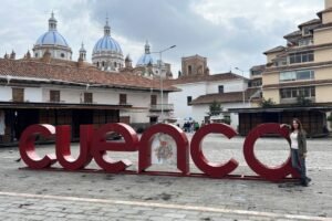 A large red “Cuenca” sign in an open plaza with a woman standing by the last letter. In the background are colonial-style buildings with terracotta roofs and the iconic blue-and-white domes of the New Cathedral of Cuenca under a cloudy sky.
