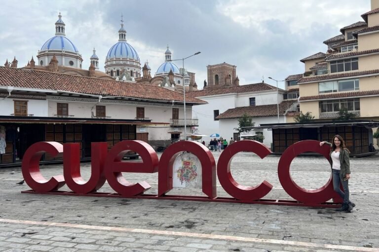 A large red “Cuenca” sign in an open plaza with a woman standing by the last letter. In the background are colonial-style buildings with terracotta roofs and the iconic blue-and-white domes of the New Cathedral of Cuenca under a cloudy sky.