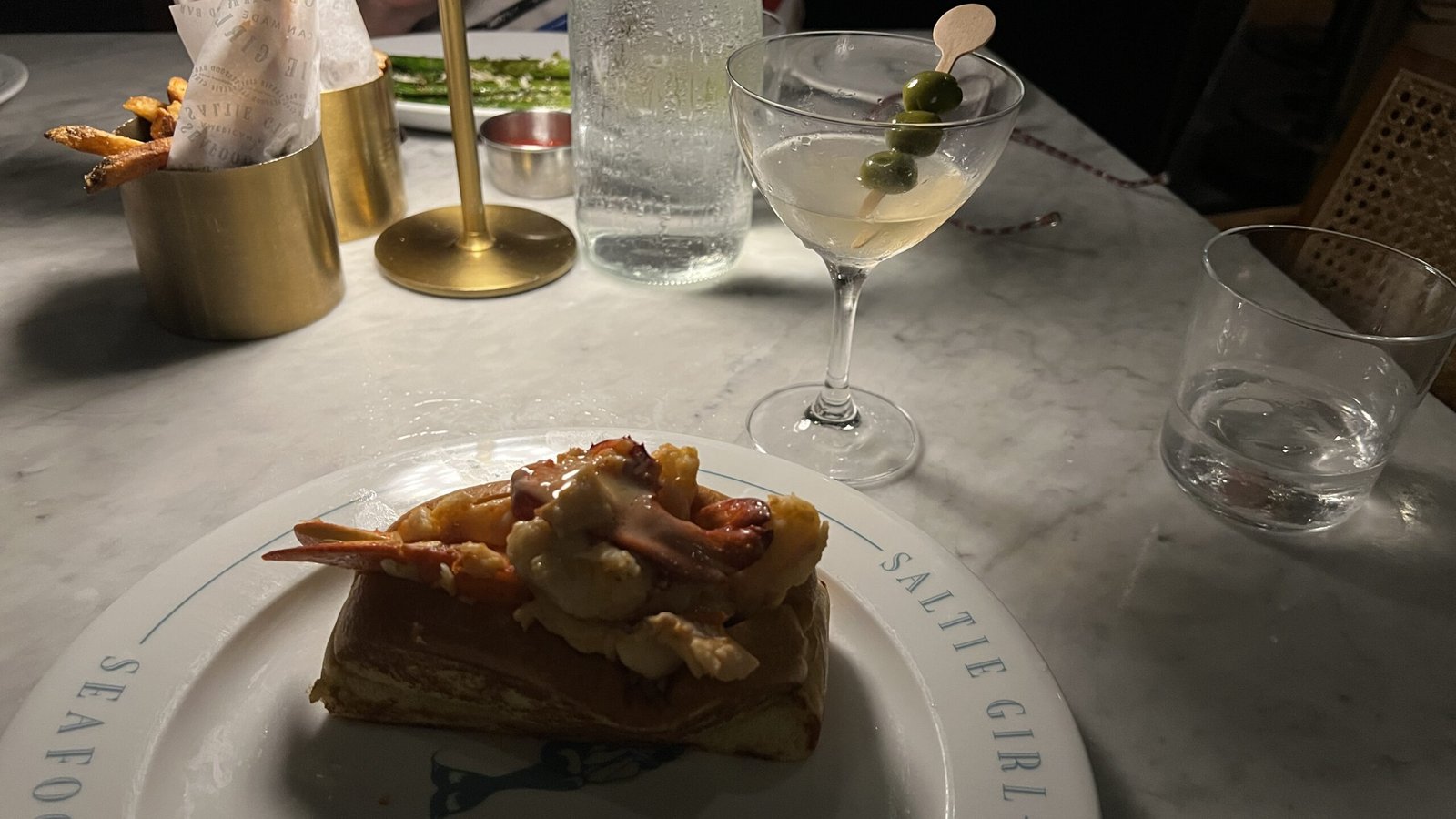 A close-up of a seafood dish from Saltie Girl—lobster served on brioche toast—alongside a martini on a white marble table.