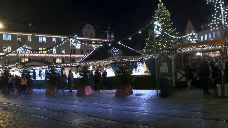 Evening view of Augsburg’s Christmas market with illuminated stalls, a decorated Christmas tree, and people walking along cobblestone streets.