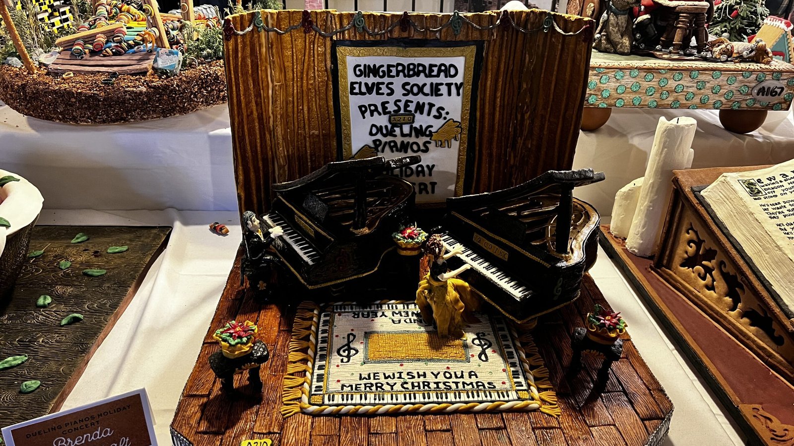 Gingerbread display featuring two grand pianos and figurines staged as a holiday performance, shown at the National Gingerbread House Competition inside the Omni Grove Park Inn & Spa.