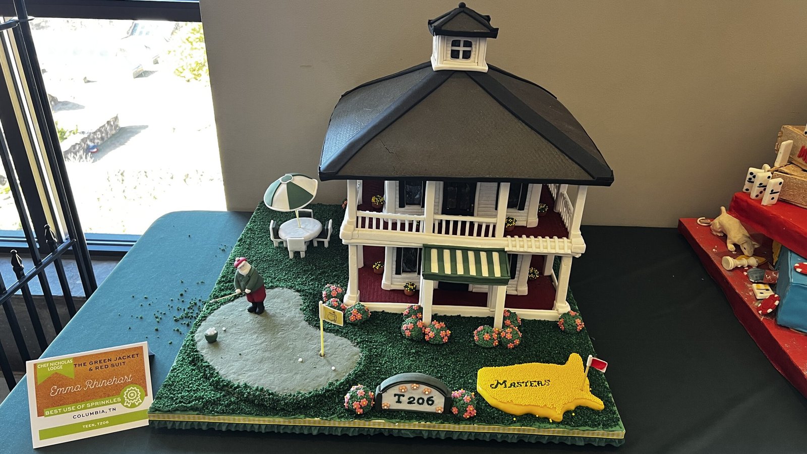 Close-up of an award card identifying a gingerbread house entry by Emma Rhinehart from Columbia, Tennessee, recognized for Best Use of Sprinkles, displayed at the National Gingerbread House Competition inside the Omni Grove Park Inn & Spa.