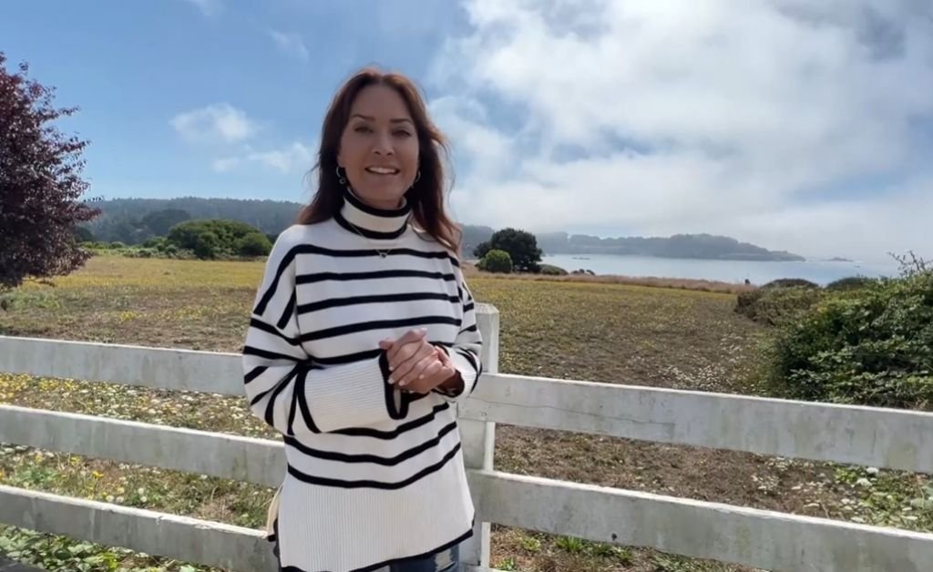 Karen stands in front of a white wooden fence overlooking a coastal meadow and fog-covered shoreline in Mendocino.