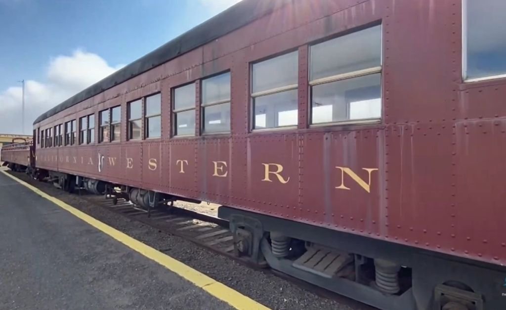 A vintage maroon California Western Railroad passenger car sits on the tracks at a station platform under a bright sky.