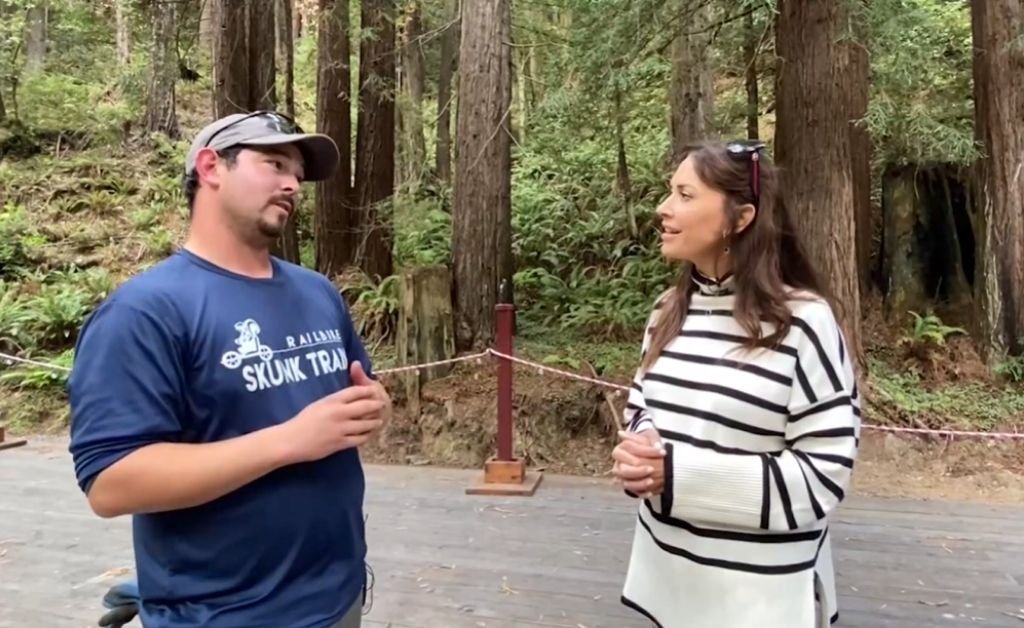Johnny, a Skunk Train guide wearing a navy Skunk Train shirt and cap, talks with Karen on a wooden platform surrounded by tall redwoods and forest greenery.