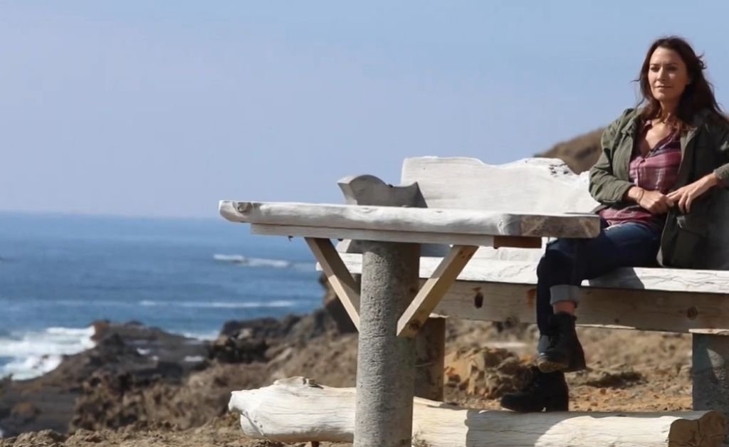 Karen sits on a driftwood bench overlooking the rocky shoreline and blue ocean on the Mendocino Coast.
