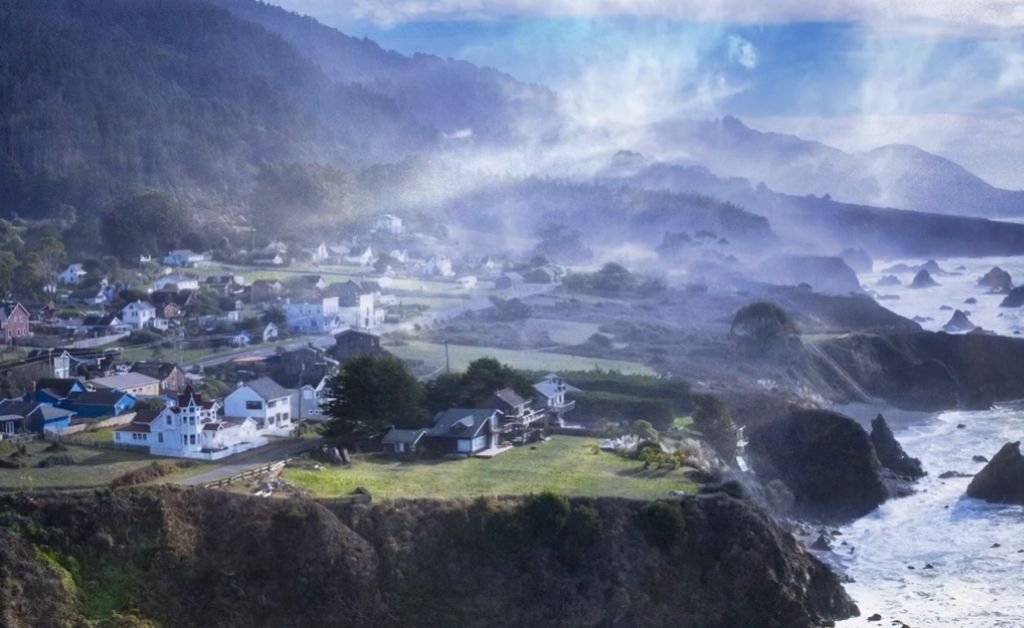 A misty aerial view of Mendocino’s coastal village, with rugged cliffs, ocean waves and forested hills rising behind the town.
