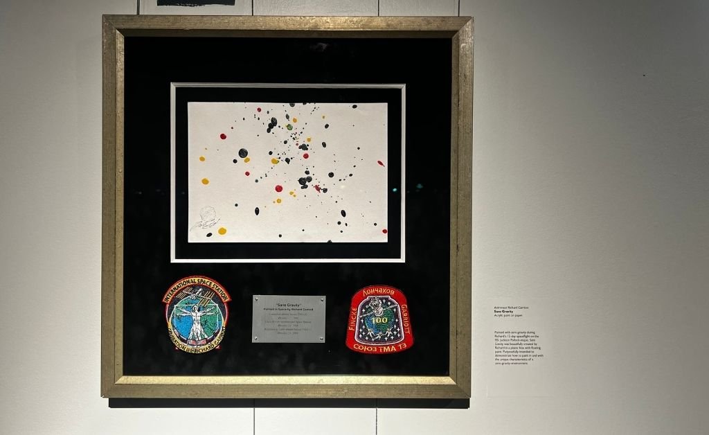 Framed artwork titled Sans Gravity by astronaut Richard Garriott, featuring splattered acrylic paint created in zero gravity aboard the International Space Station, displayed with mission patches.