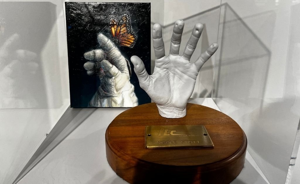 Cast of astronaut Nicole Stott’s hand mounted on a wooden base by ILC Dover, displayed alongside a painting of an astronaut glove reaching toward a butterfly.