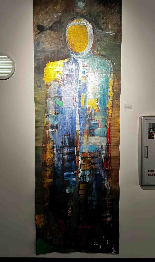 Large vertical abstract painting depicting a human figure in a spacesuit, rendered in layered textures of blue, yellow, and earth tones, displayed at Orlando Science Center.